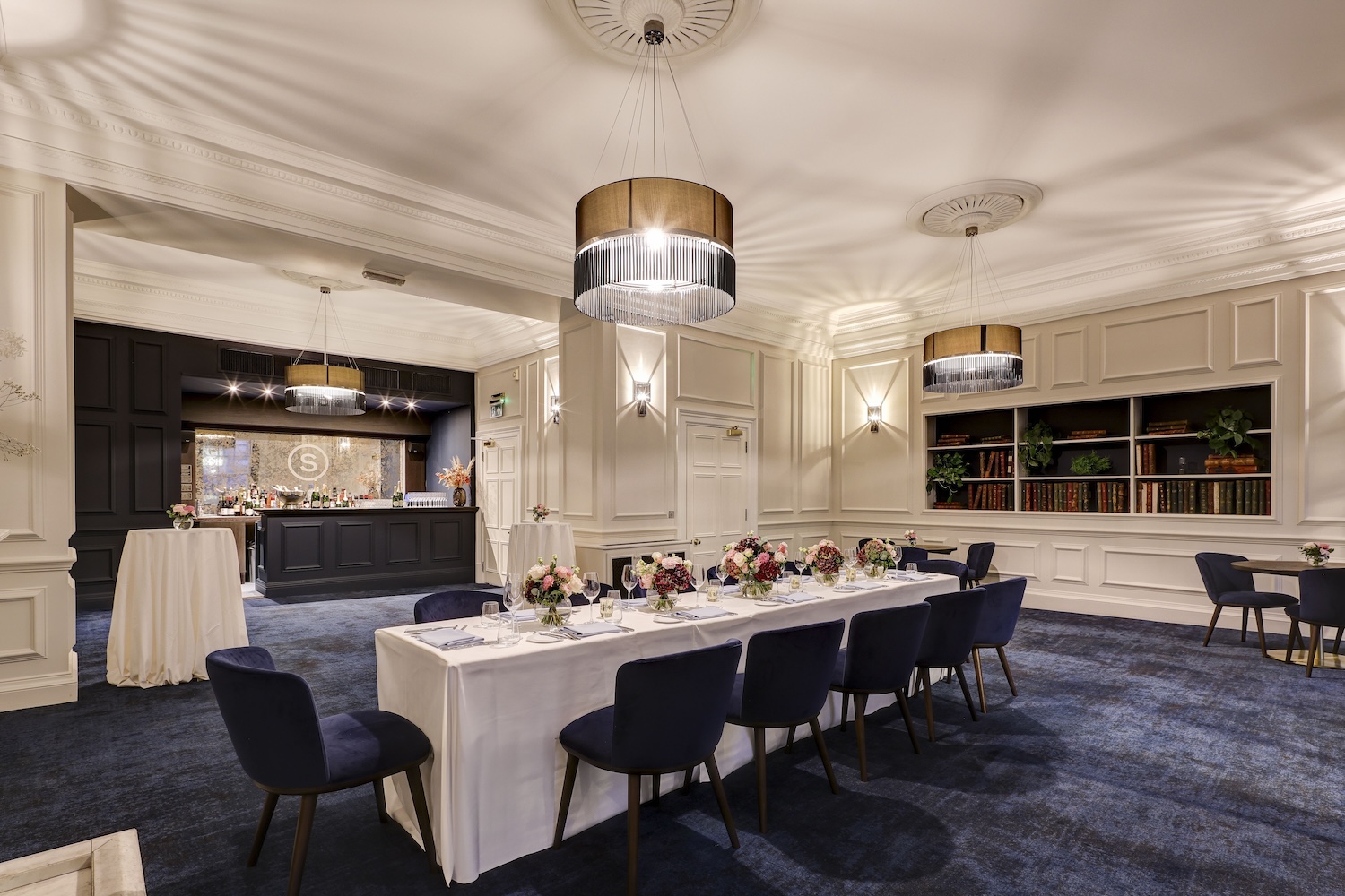 A well lit grand room with high ceilings and a royal blue carpet. In the centre of the room is a laid table, featuring a white tablecloth, napkins and wine glasses. In the background of the room is a bar with champagne bottles laid out.