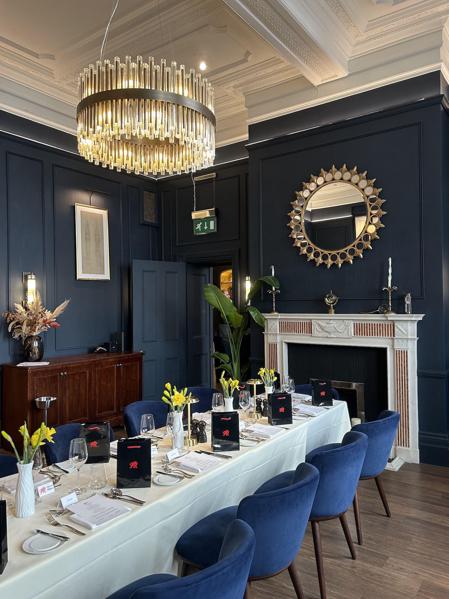 A medium sized room with dark hard wood flooring, high ceilings and royal blue painted walls. There is a well laid table with flowers, menus, napkins and cutlery on it. The room also features a fireplace and a circular mirror above it.
